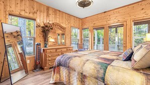 A wooden cabin bedroom with a large bed, dresser, and a mirror. There are multiple windows providing views of the surrounding forest.