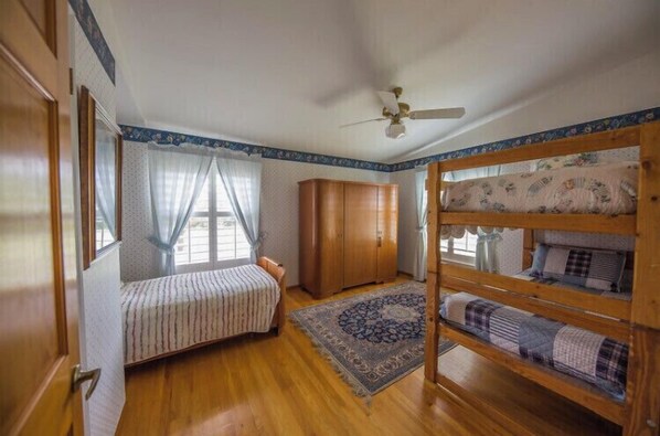 Quarto com cama, guarda-roupa, janela com cortinas, ventoinha no teto e tapete estampado.