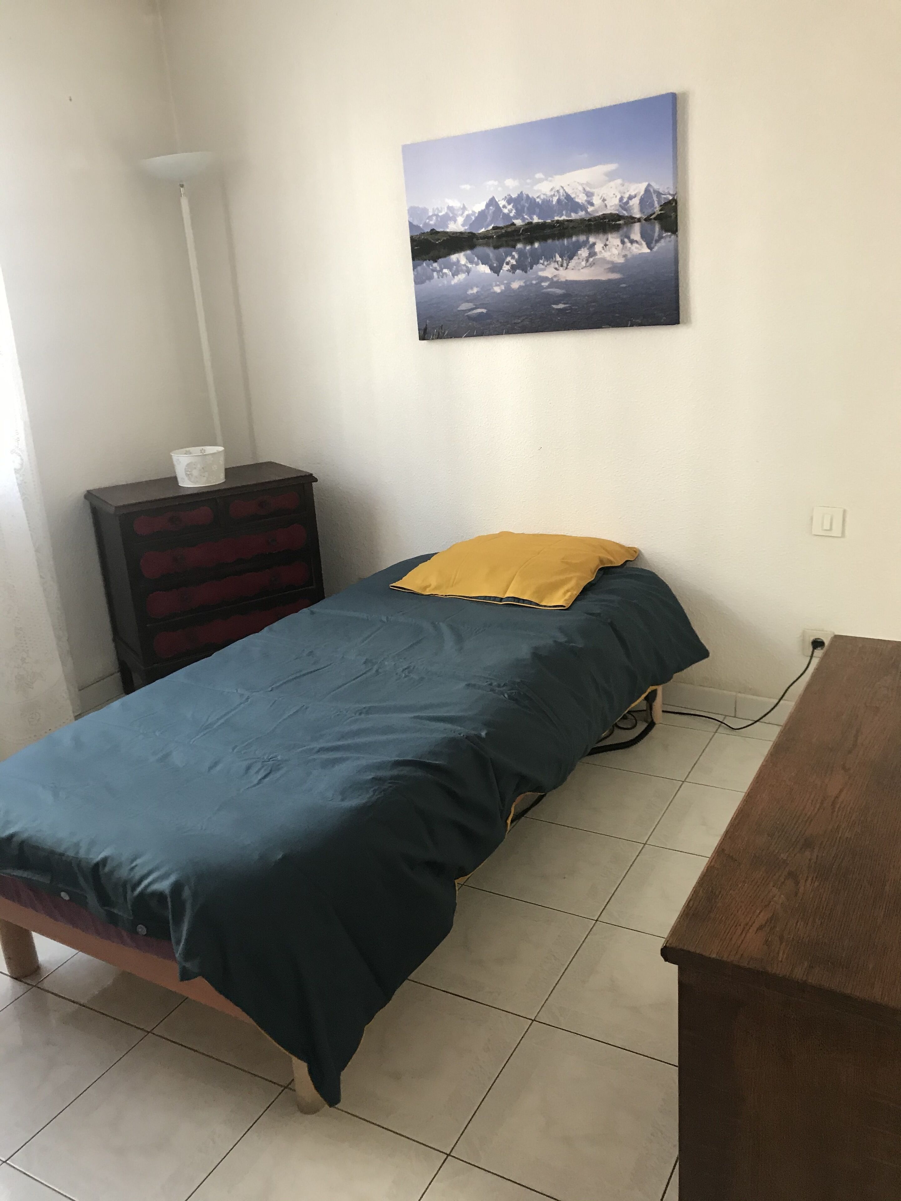 A small, simple room with a bed, a dresser, and a painting on the wall.
