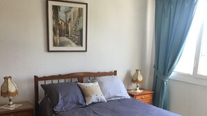 Un dormitorio con una cama de madera, una mesita de noche con lámpara, un cuadro en la pared y una ventana con cortinas.