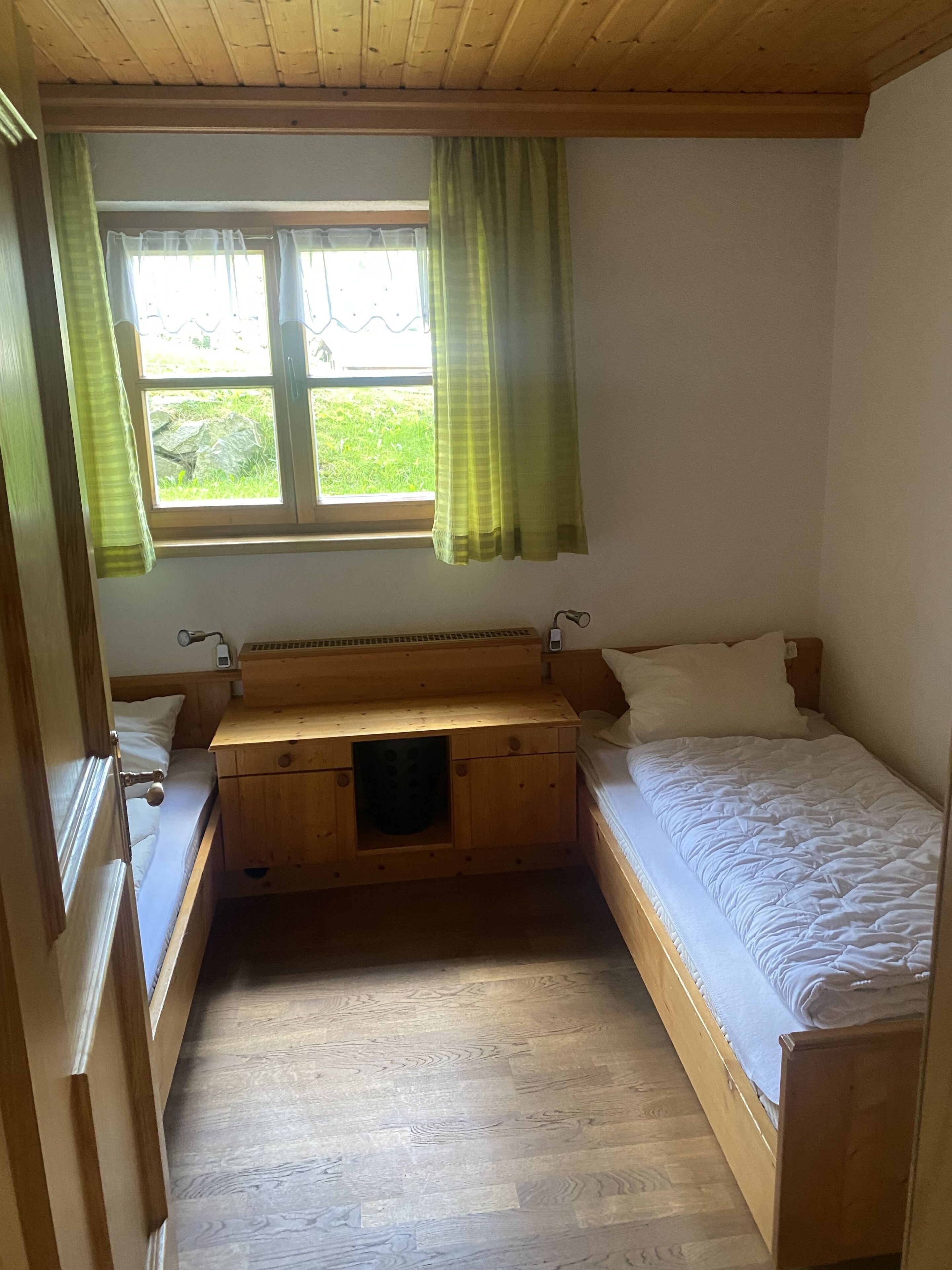 Ein kleiner, kompakter Raum mit zwei Holzbett, einem Fenster mit grünen Vorhängen und einem Holztisch mit Lampe.