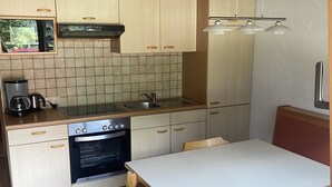 Cocina compacta con armarios de madera, horno integrado, microondas y cafetera. Cuenta con una mesa de comedor con sillas y un banco.