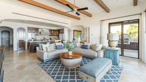 A spacious living room with a large round coffee table, a sofa, and a blue ottoman. There is a kitchen in the background with a large window. - 3 BD Oceanview Penthouse with infinity pool and jacuzzi (San Jose del Cabo)