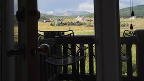 Uitzicht vanuit een kamer met een deur naar buiten, met een balkon met stoelen en een tafel, uitkijkend over een landelijk landschap met huizen en bergen.