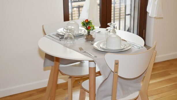 Una mesa de comedor lista con platos blancos, cubiertos y un pequeño florero con una flor. Hay dos sillas con patas de madera y tapicería blanca.