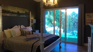 Una cama con dosel y cuatro columnas, una araña y vistas a una piscina y jardín a través de amplios ventanales correderas.