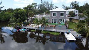 Une maison à deux étages avec une jetée qui s’avance sur une étendue d’eau calme, le tout entouré d’une végétation luxuriante.