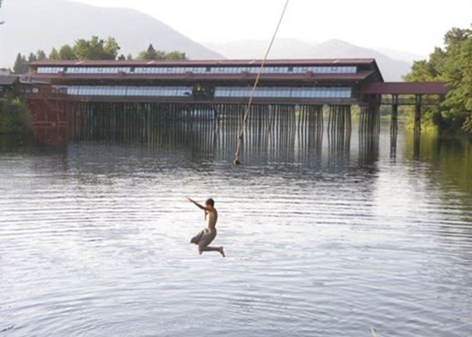 A person swinging on a rope over water with a building in the background.