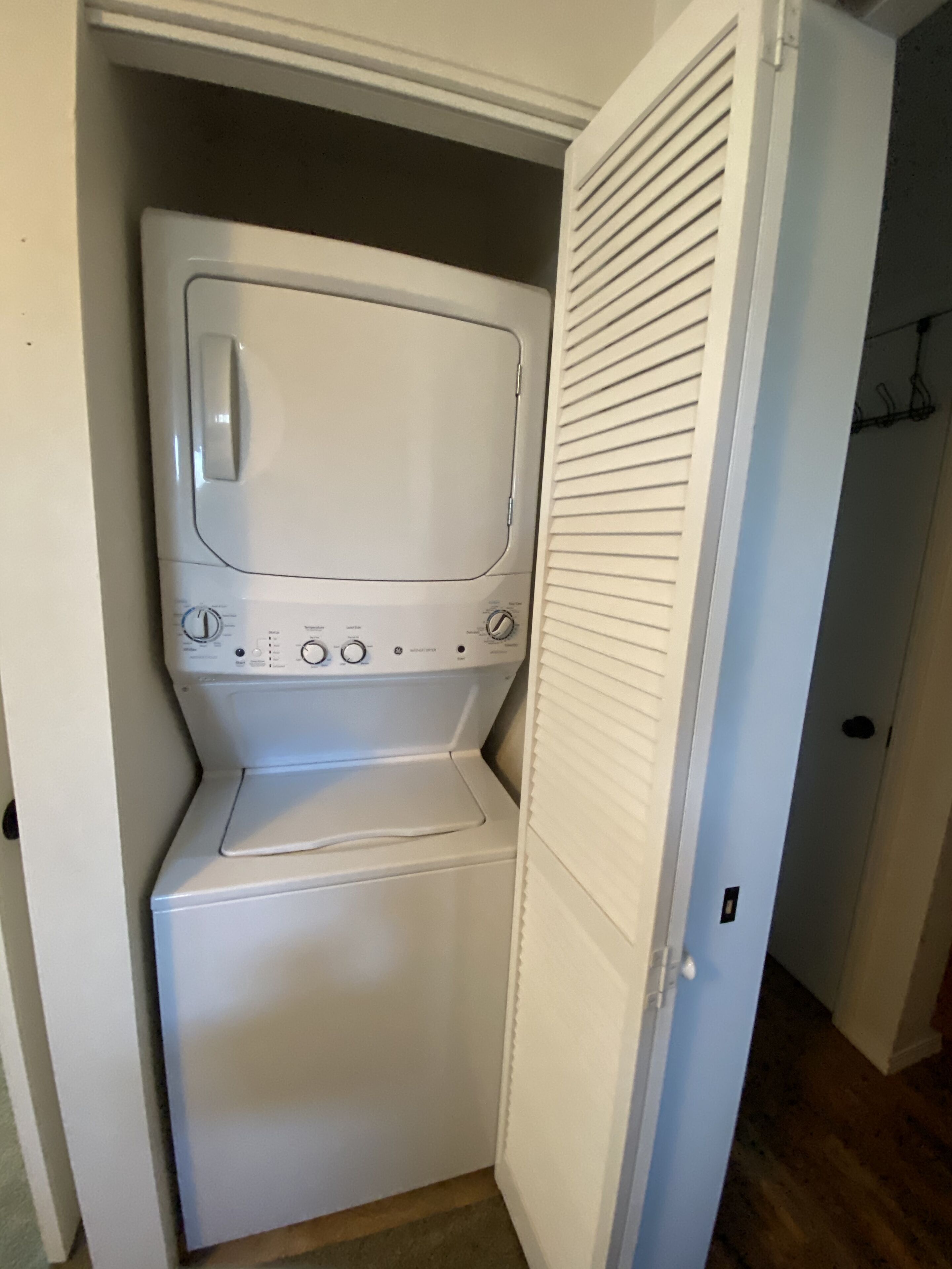 A compact laundry room with a front-loading washer and dryer, a door with a louvered window, and a wooden floor.