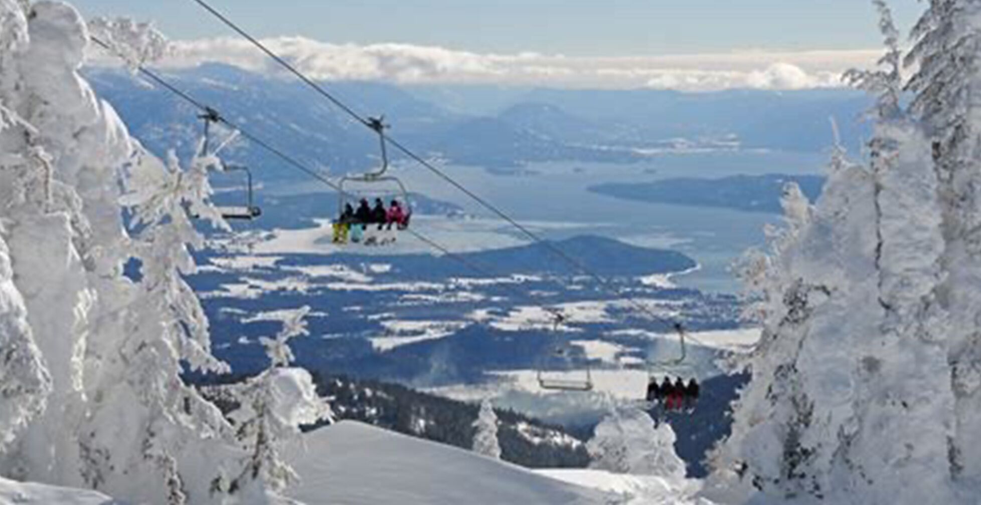 A ski lift with passengers, surrounded by snow-covered mountains and a lake.