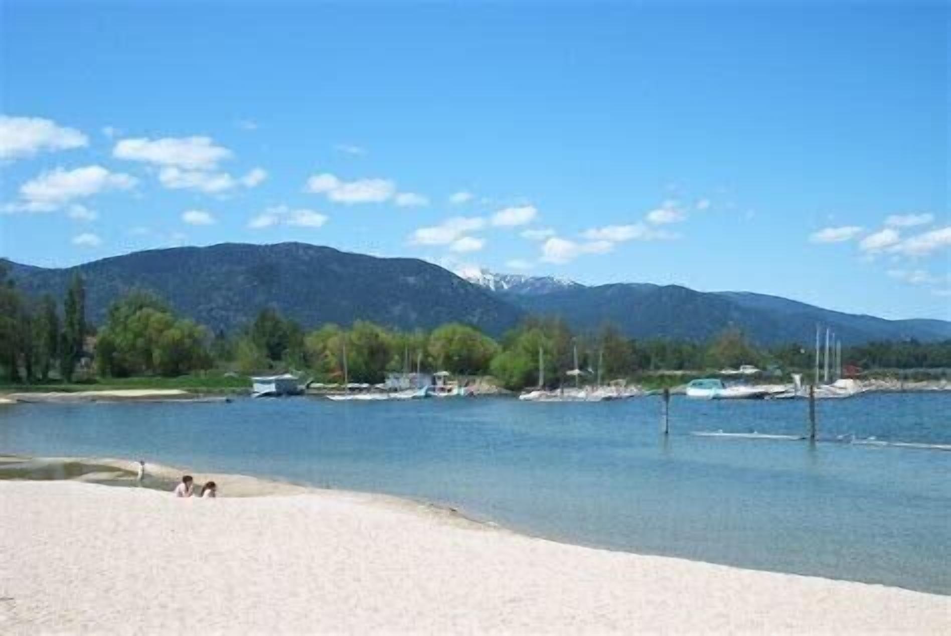 A lakeside view with boats, a sandy beach, and mountains in the background.