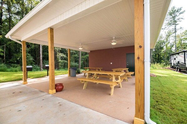 A covered picnic area with wooden picnic tables and a concrete floor, surrounded by greenery. - WoodSpring Suites Atlanta Airport (Atlanta)