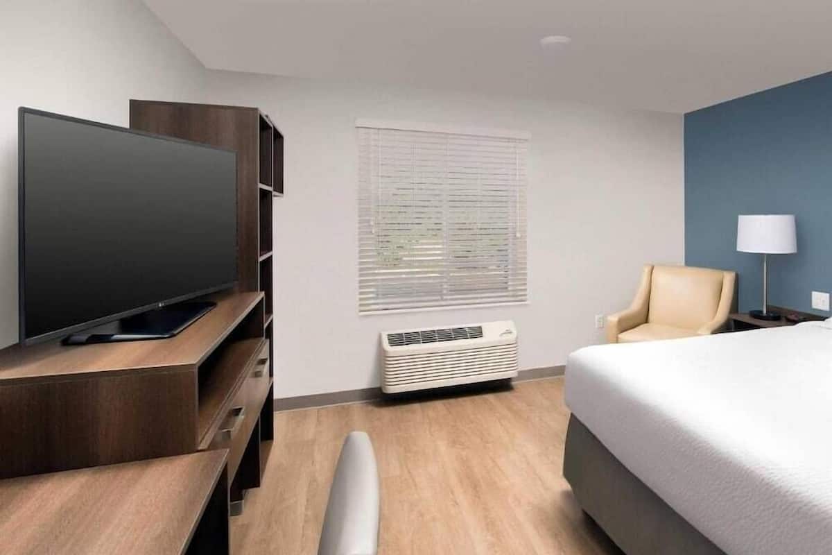 A hotel room with a flat-screen TV, a desk, a chair, a bed, a lamp, and a window with blinds.