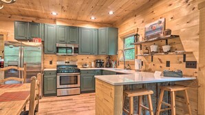 A wooden cabin kitchen with green cabinets, stainless steel appliances, a central island, and a window overlooking greenery. - Peaceful Cabin on 3 Private Acres: Deck & Fire Pit (Blairsville)