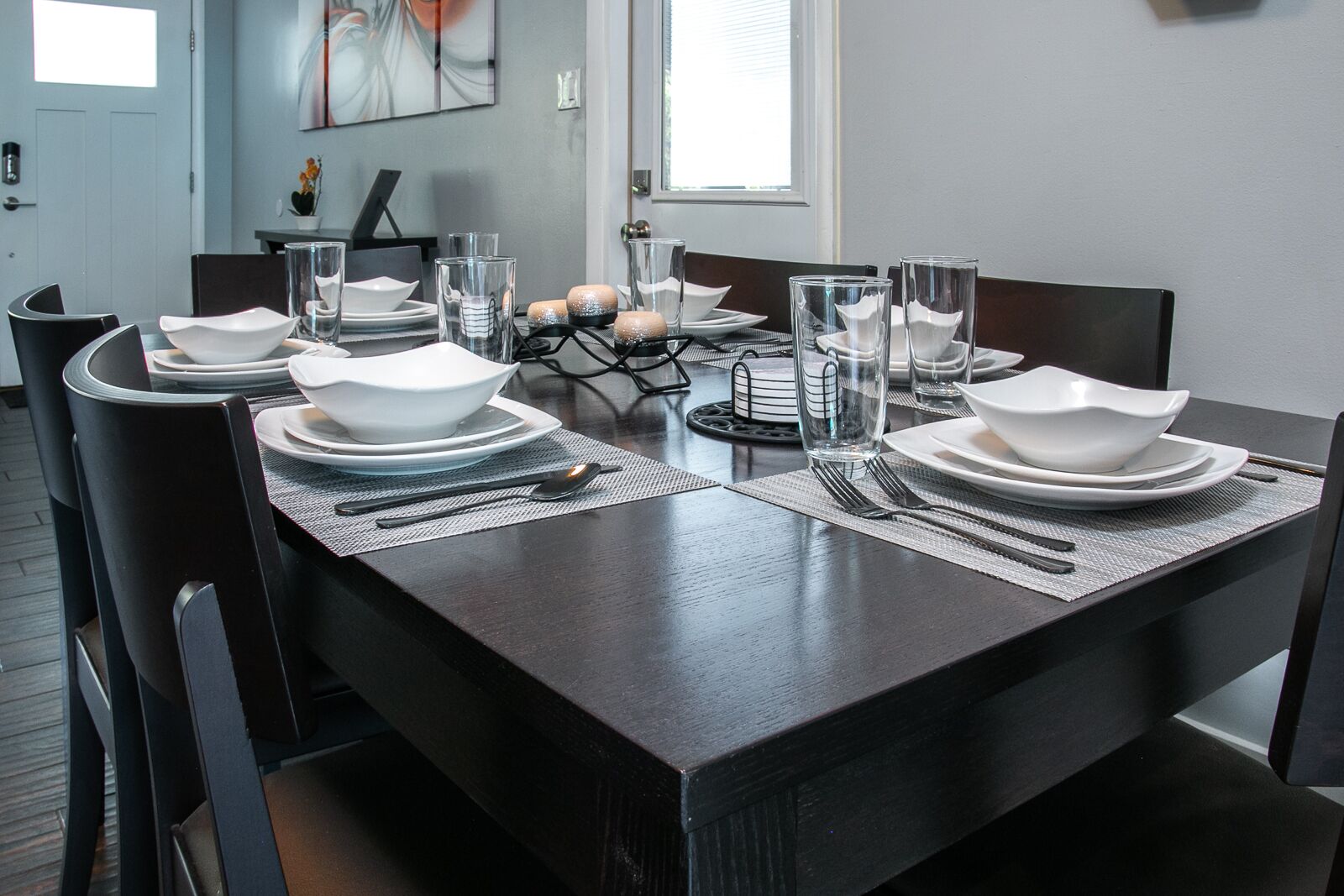 Una mesa de comedor dispuesta con platos, tazones, vasos y cubiertos blancos, de diseño moderno y con vista al salón.