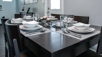 Una mesa de comedor dispuesta con platos, tazones, vasos y cubiertos blancos, de diseño moderno y con vista al salón.