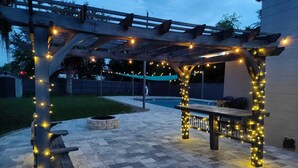 Un patio exterior con una pérgola, luces colgantes, un pozo de fuego y una piscina al fondo.