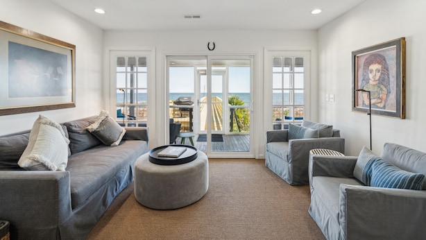 Ein großzügiges Wohnzimmer mit einem großen grauen Sofa, einem runden Hocker und Meerblick durch Glastüren.