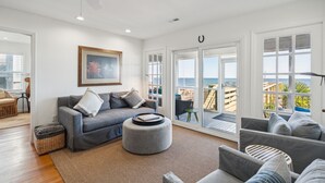 Ein modernes Wohnzimmer mit grauem Sofa, Hocker und Sesseln, einem Deckenventilator und großen Fenstern mit Blick auf das Meer.