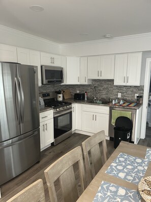 A modern kitchen with white cabinets, stainless steel appliances, and a wooden dining table. - Beach Summer House (Ventnor)