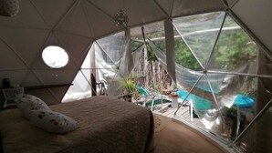 Um quarto tipo tenda com uma cama, uma mesinha e uma cadeira. Há janelas e vista para o verde lá fora.