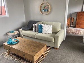 Un salon avec un canapé vert clair, une table basse en bois, une petite table d’appoint ronde sur laquelle se trouvent un bol bleu et un pichet, ainsi qu’une étagère murale ornée d’objets décoratifs.