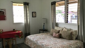 Un dormitorio con cama, una mesa con mantel rojo, una planta pequeña, un marco de fotos y una ventana con cortinas.