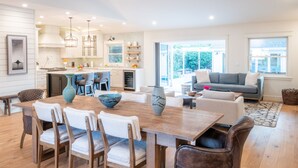 Un amplio salón con una mesa de comedor de madera, sillas blancas, un sofá gris y una cocina al fondo.