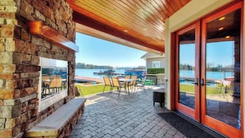 Un patio esterno con pavimento in pietra, soffitto in legno e parete in pietra. Si vedono un tavolo e sedie, e una vista su un lago con barche.