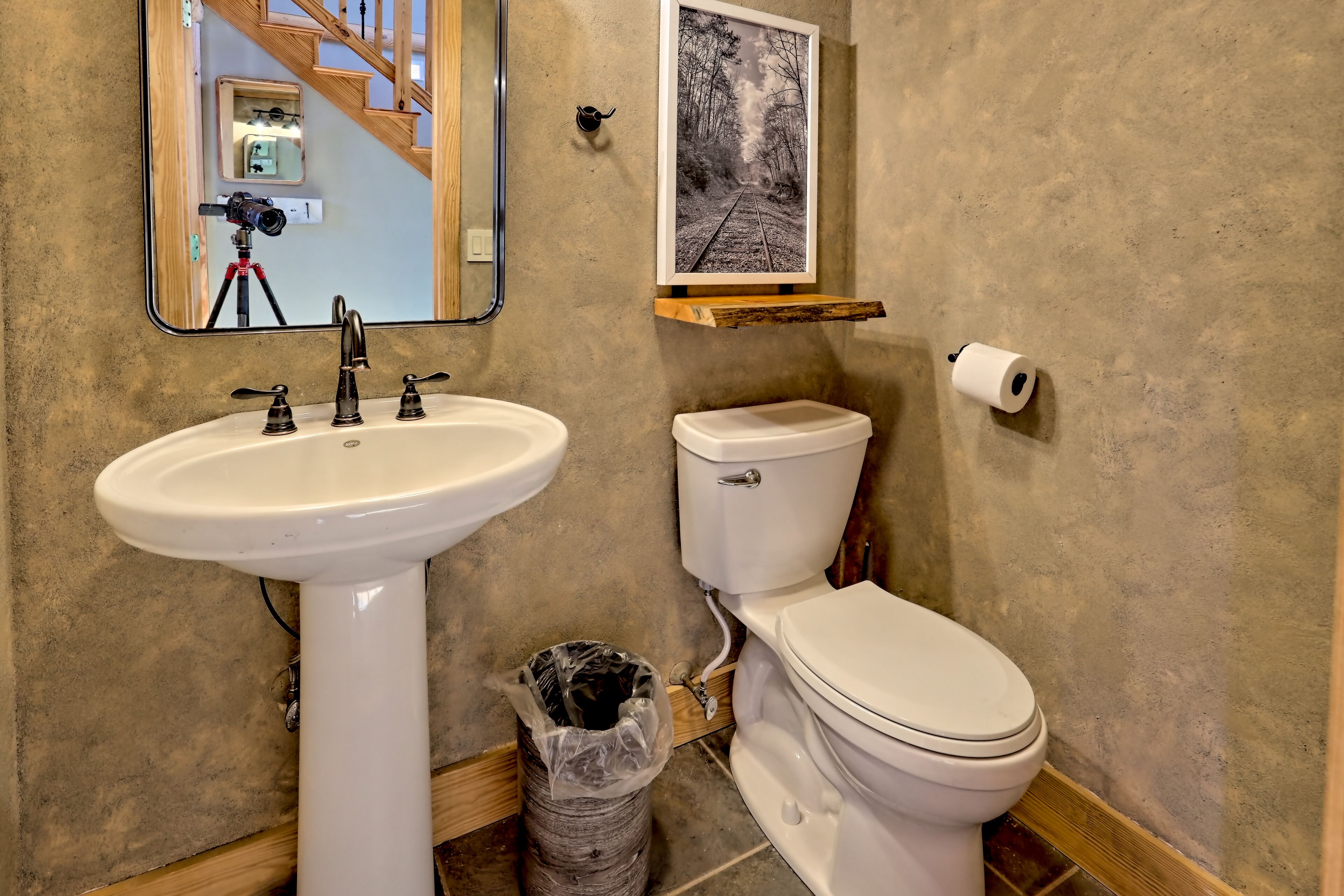 A bathroom with a white sink, a toilet, a wooden shelf with a framed picture, and a trash can.