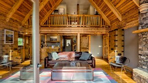Een ruime woonkamer met een hoge plafond, houten balken, een leren bank, een bijzettafeltje en een stenen open haard.