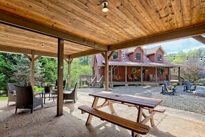 Un espacio al aire libre cubierto con mesas y sillas de picnic, una terraza de madera y una vista de una casa grande al fondo.