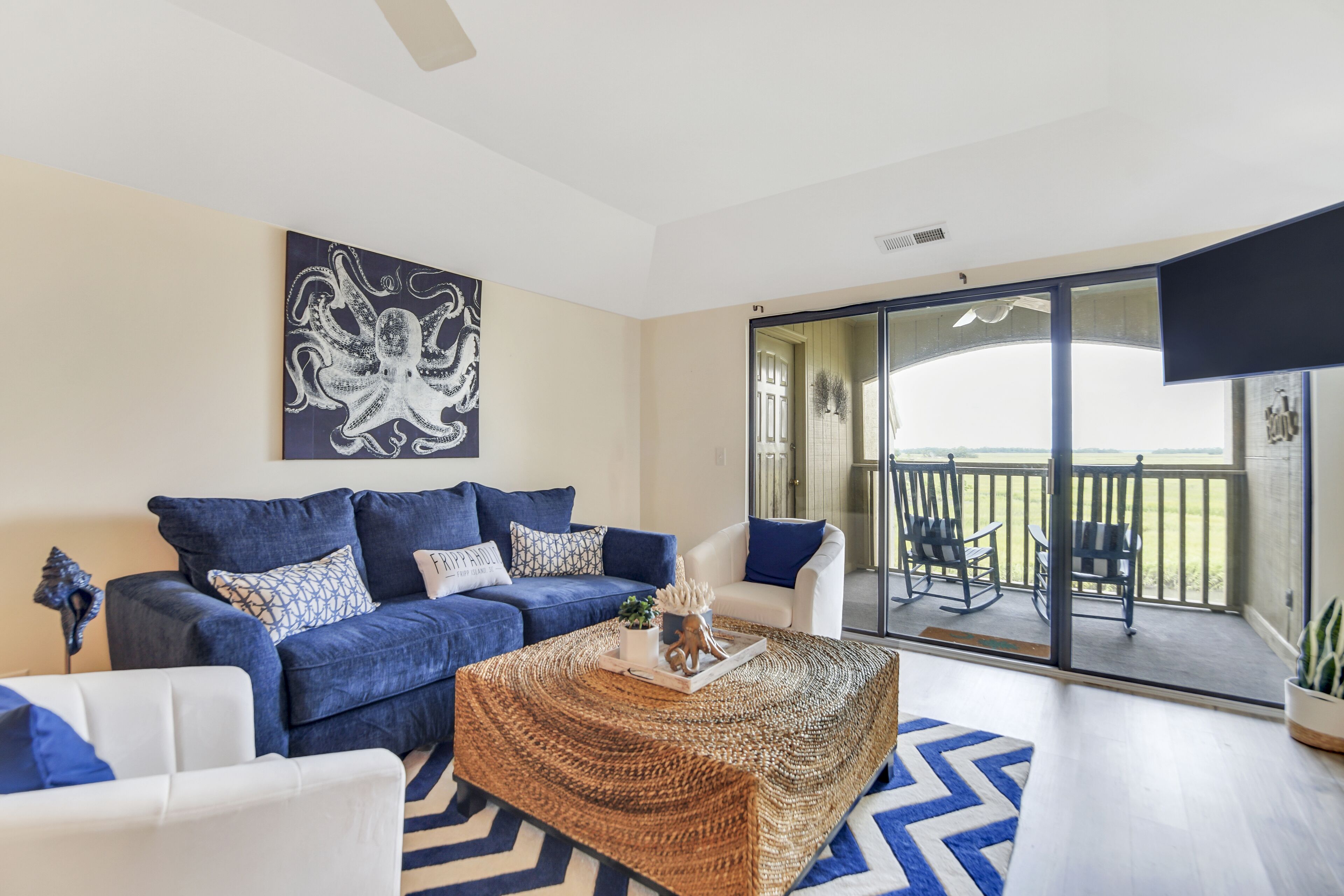 A living room with a blue sofa, white armchairs, a woven coffee table, and a large painting of an octopus.
