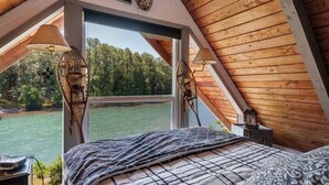 Ein Schlafzimmer mit großem Fenster mit Blick auf ein Gewässer und einen Wald. Es gibt ein Bett mit gemustertem Überwurf und zwei Wandlampen.