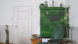 Une pièce avec une cheminée recouverte de carreaux verts, une chaise en bois, une petite table en bois ornée d’un vase et un lampadaire.