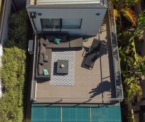 An aerial view of a rooftop patio with a pool, a seating area with a patterned rug, and surrounding greenery.