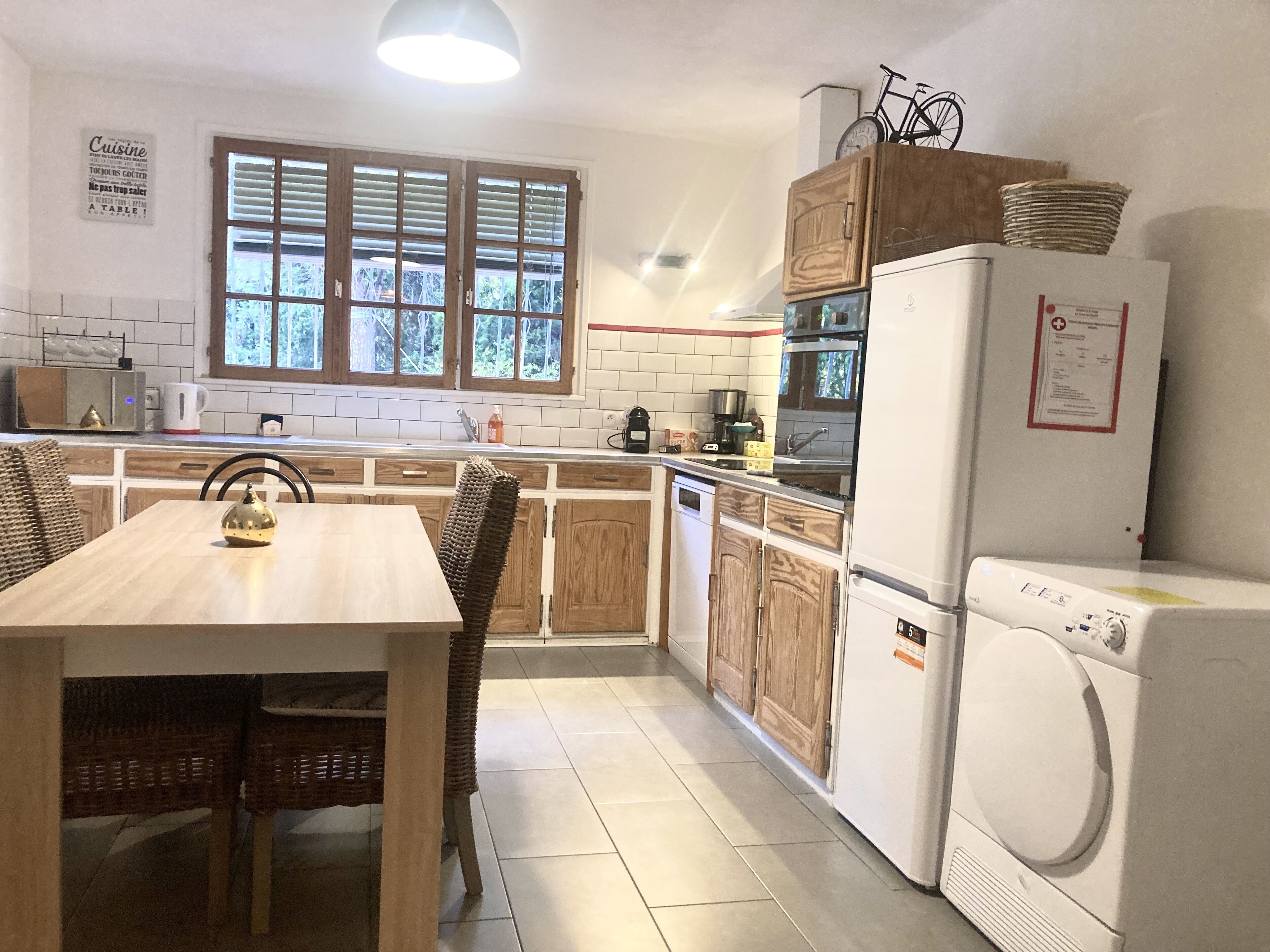 Une cuisine avec des meubles en bois, une table à manger avec des chaises, un lave-linge et une fenêtre avec des stores.