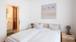 A hotel room with a bed, two bedside tables with lamps, a nightstand, a wooden door, and a wall-mounted painting.