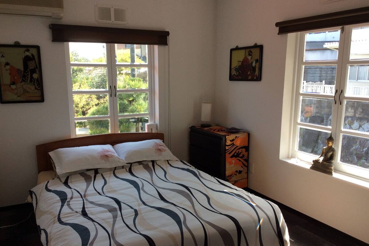 A bedroom with a bed, two windows, a nightstand, and a Buddha statue.