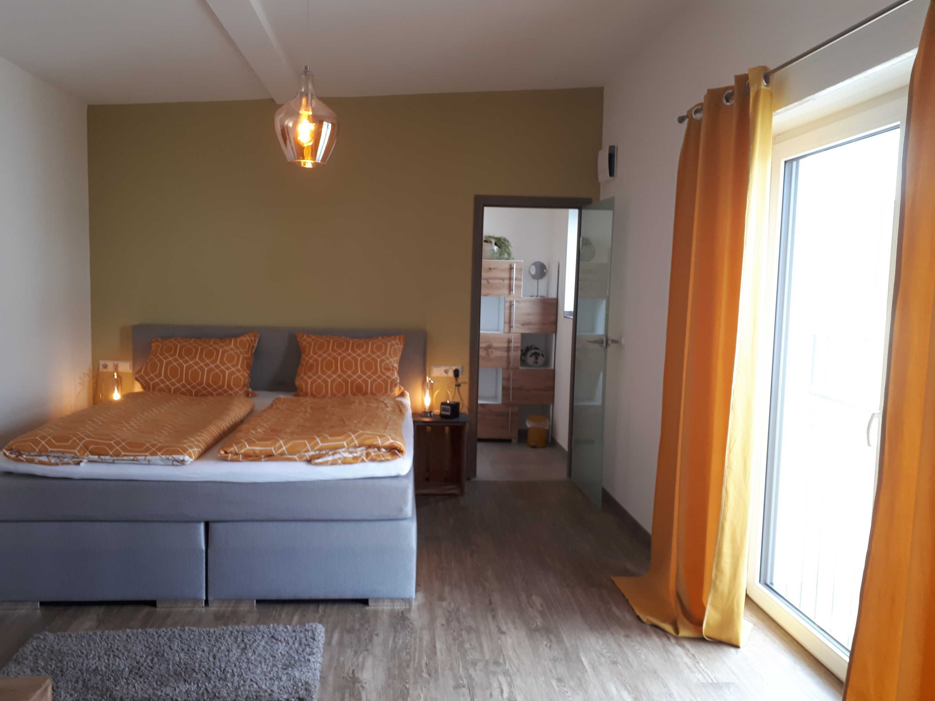 A bedroom with a bed, bedside tables, a hanging light fixture, a sliding glass door, and a view of a hallway.