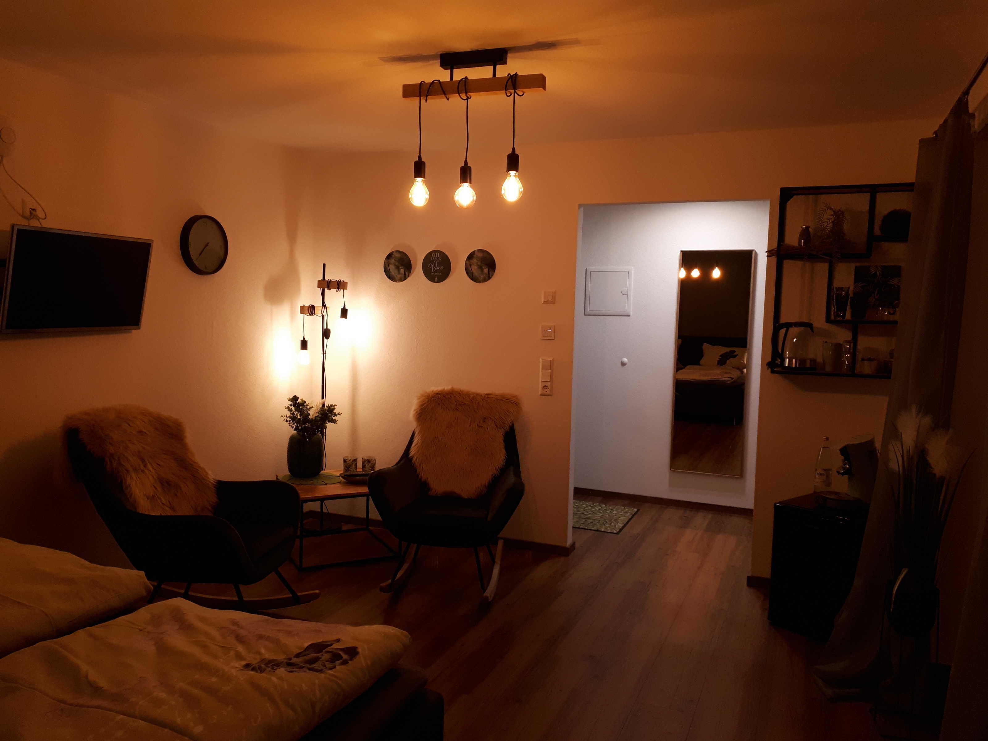 A cozy living room with a sofa, two armchairs, a coffee table, and a floor lamp. There is a door leading to another room, and a shelf with various items on the right.