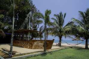 Escena de playa con una estructura de barco de madera, palmeras y una hamaca.