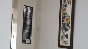 A hallway with a white wall, a black-framed painting of flowers, a potted plant, and a window with blinds. - Flip Flops Hostel (Pasay)