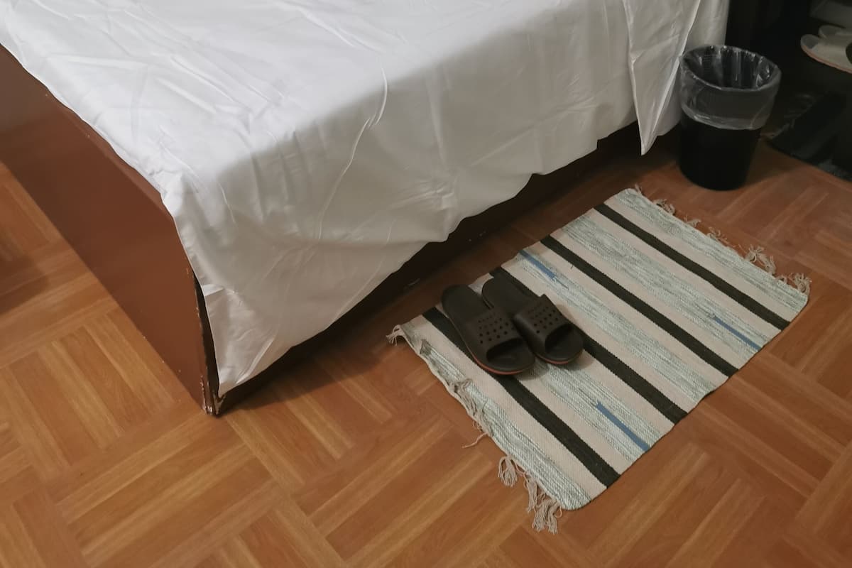 En snyggt bäddad säng med vita lakan och kuddar, en randig matta, en papperskorg och ett litet bord med en lampa.