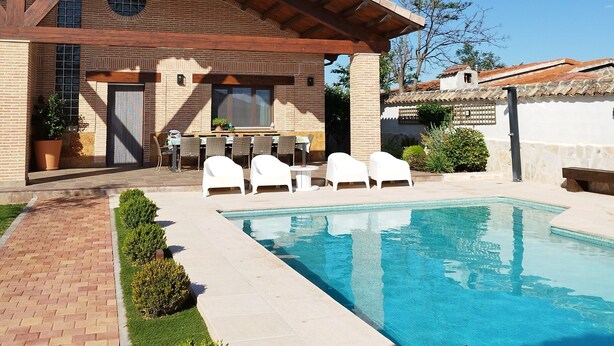 Un espace aménagé au bord de la piscine, avec un bâtiment en briques, des sièges extérieurs et un ciel d’un bleu limpide.