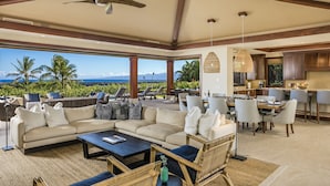 A spacious living area with a large sofa, dining table, and chairs, overlooking a scenic view with palm trees and mountains. - Luxury 5BR Kulanakauhale Estate 3558 at Hualalai – Infinity Pool & Ocean Views (Kailua Kona)