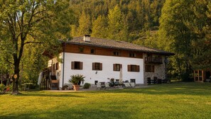 A two-story building with a white facade and wooden shutters, surrounded by a well-maintained lawn and dense forest.