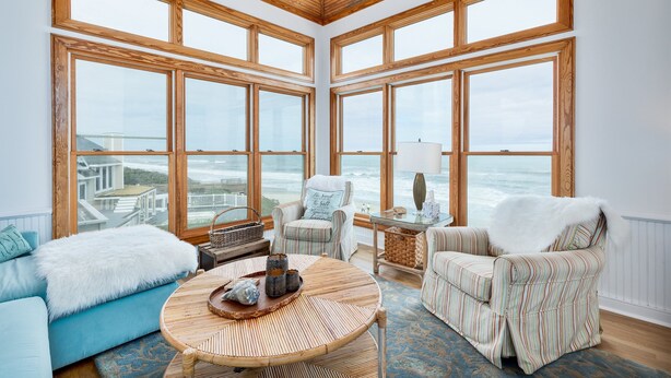 A spacious room with large windows, a blue sofa, striped armchairs, and a round wooden table. - 2065 - Sweet Home Carolina - Oceanfront Escape w/ Pool, Hot Tub & Views (Corolla)