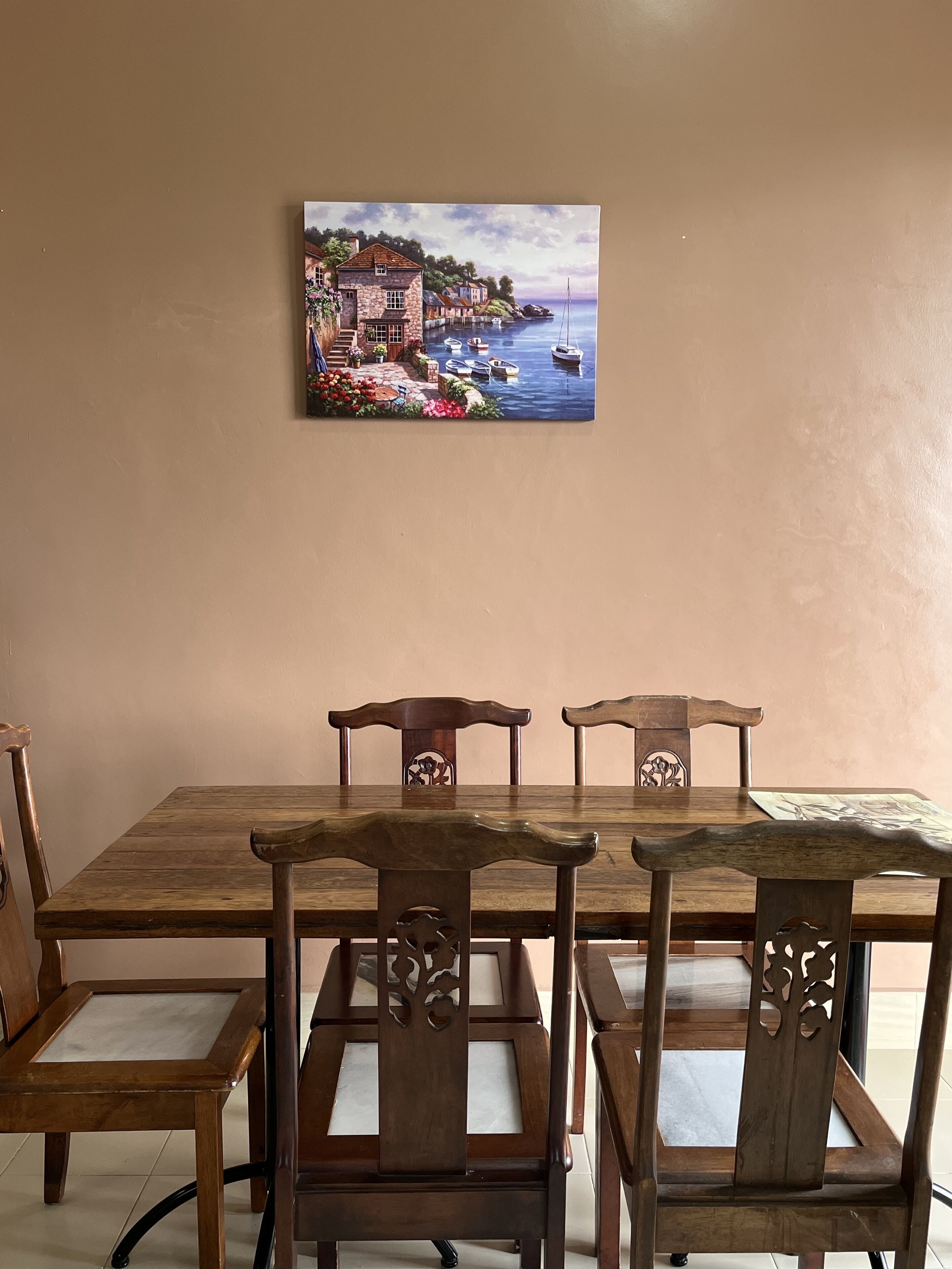 A dining table with six wooden chairs, a painting of a coastal scene on the wall, and a tiled floor.