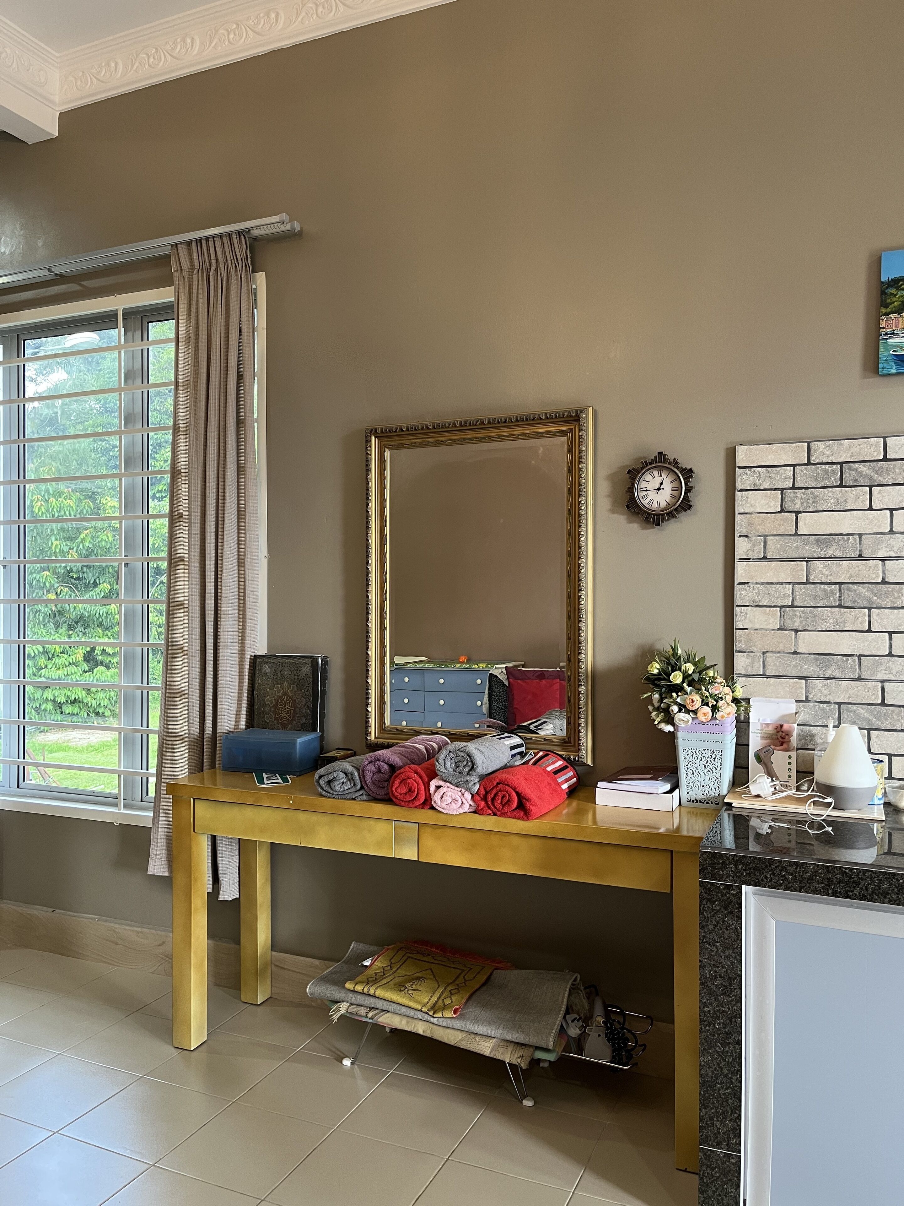 A room with a wooden desk, a mirror, a clock, a brick-patterned wall, and a window with curtains.